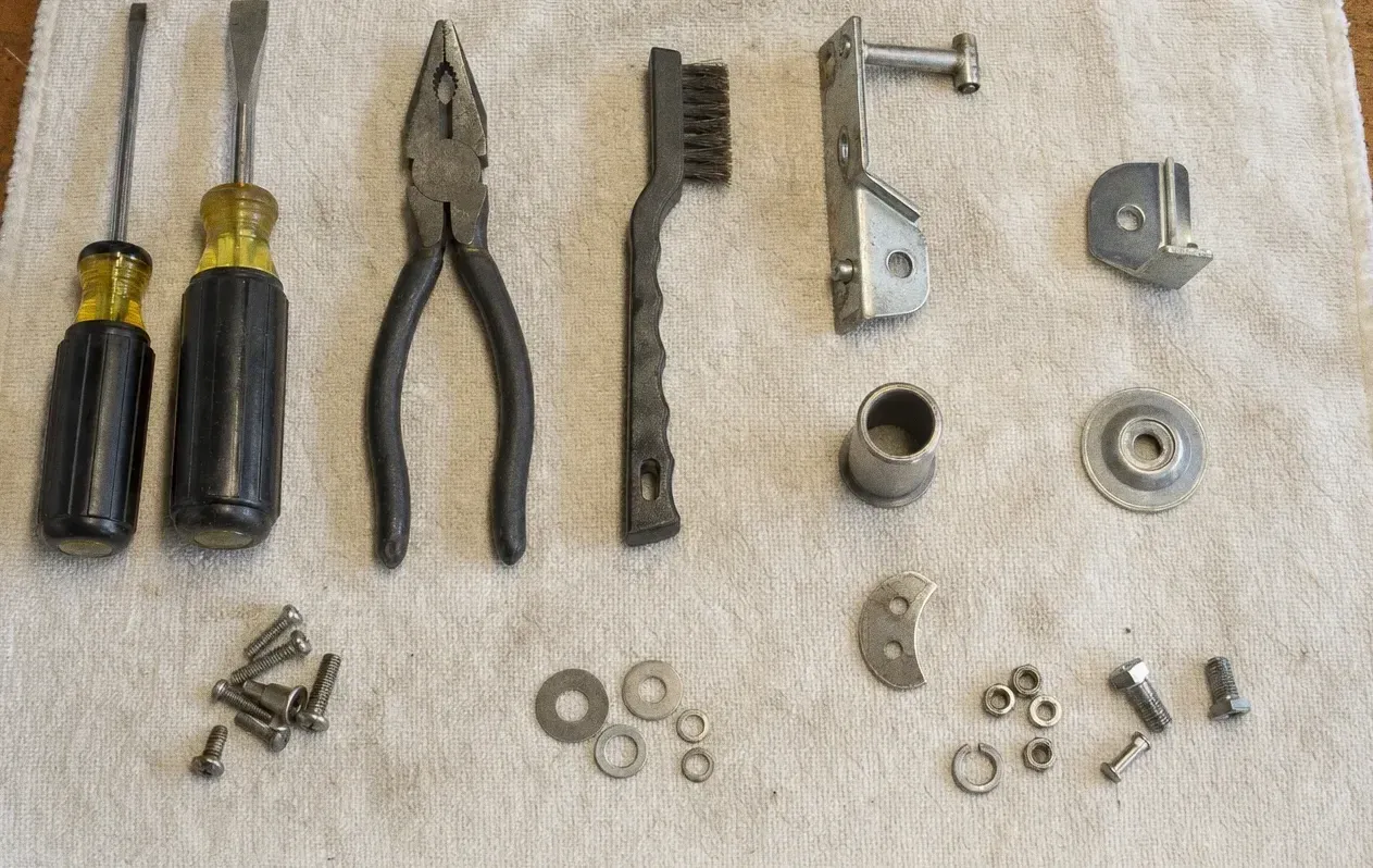 Screwdrivers, pliers and a wire brush laid out with a hinge arm, a bronze bushing, a sealed bearing, washers and bolts