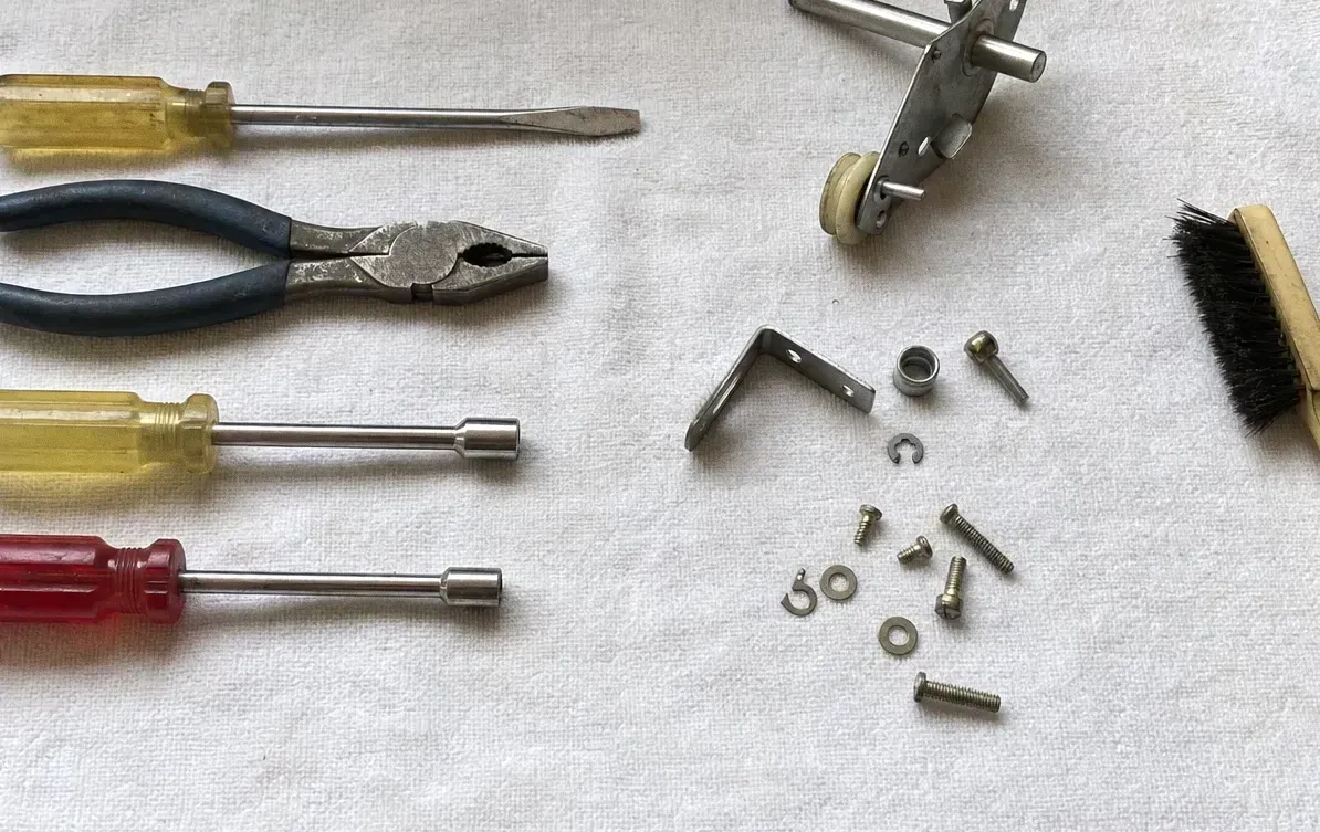 Lineman’s pliers, two nut drivers and a screwdriver laid out with a hinge roller assembly, loose screws and washers on a towel